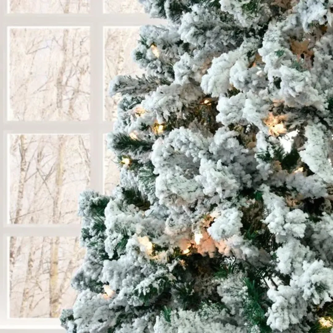 Frosted Christmas tree with lights in front of a window with a snowy landscape view.